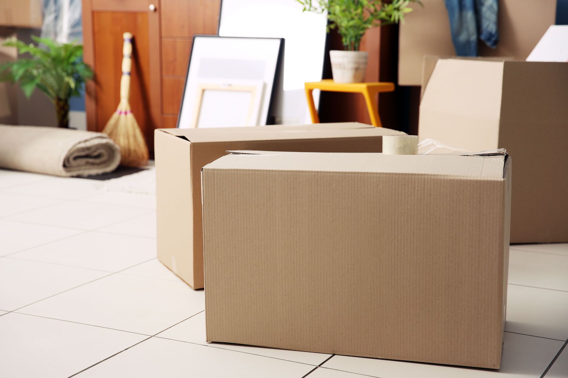 Umzugssituation mit Kartons und Möbeln. Im Vordergrund stehen zwei große Umzugskartons auf gefliestem Boden. Im Hintergrund sind ein zusammengerollter Teppich, ein Besen, Bilderrahmen und Pflanzen zu sehen.